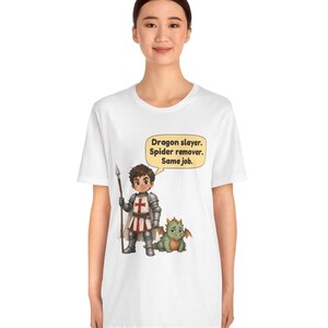 K&ouml;nnte beinhalten: Ein wei&szlig;es T-Shirt mit einer Grafik eines Ritters in silberner R&uuml;stung, der einen Speer neben einem kleinen gr&uuml;nen Drachen h&auml;lt. Eine Sprechblase dar&uuml;ber enth&auml;lt den Text: Dragon slayer. Spider remover. Same job.