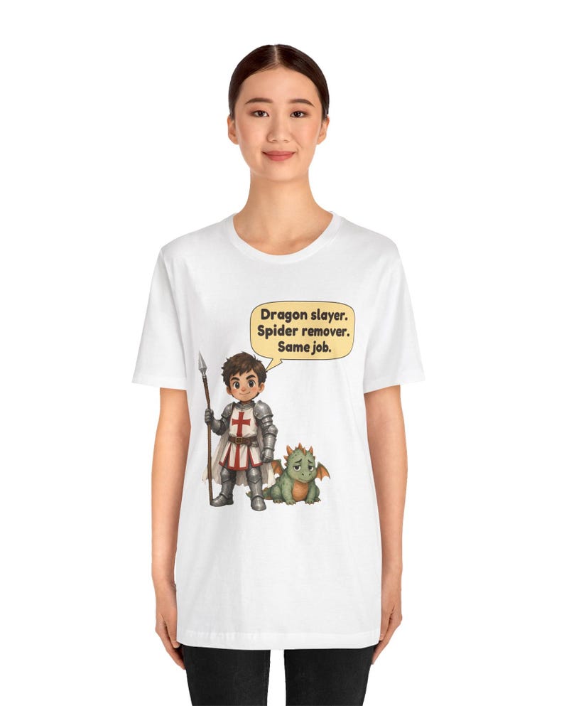 K&ouml;nnte beinhalten: Ein wei&szlig;es T-Shirt mit einer Grafik eines Ritters in silberner R&uuml;stung, der einen Speer neben einem kleinen gr&uuml;nen Drachen h&auml;lt. Eine Sprechblase dar&uuml;ber enth&auml;lt den Text: Dragon slayer. Spider remover. Same job.