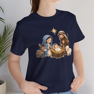 May include: A navy blue t-shirt featuring a graphic illustration of a nativity scene. The design includes a central infant in a manger, a figure in a blue head covering, a figure in a brown robe, a donkey, a lamb, and a gold star above.