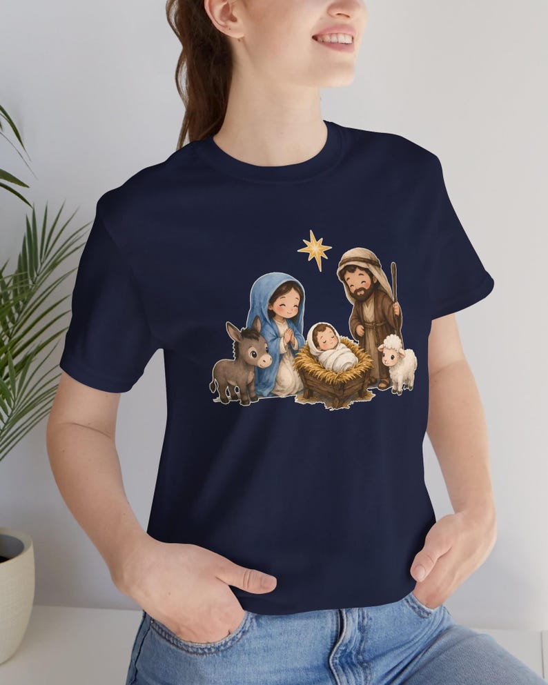 May include: A navy blue t-shirt featuring a graphic illustration of a nativity scene. The design includes a central infant in a manger, a figure in a blue head covering, a figure in a brown robe, a donkey, a lamb, and a gold star above.