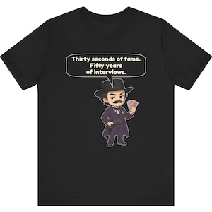 Pu&ograve; includere: Una t-shirt nera con un'illustrazione in stile cartoon di un personaggio con i baffi, cappotto scuro e cappello che tiene in mano delle carte da gioco. Un fumetto sopra il personaggio contiene il testo: Thirty seconds of fame. Fifty years of interviews.