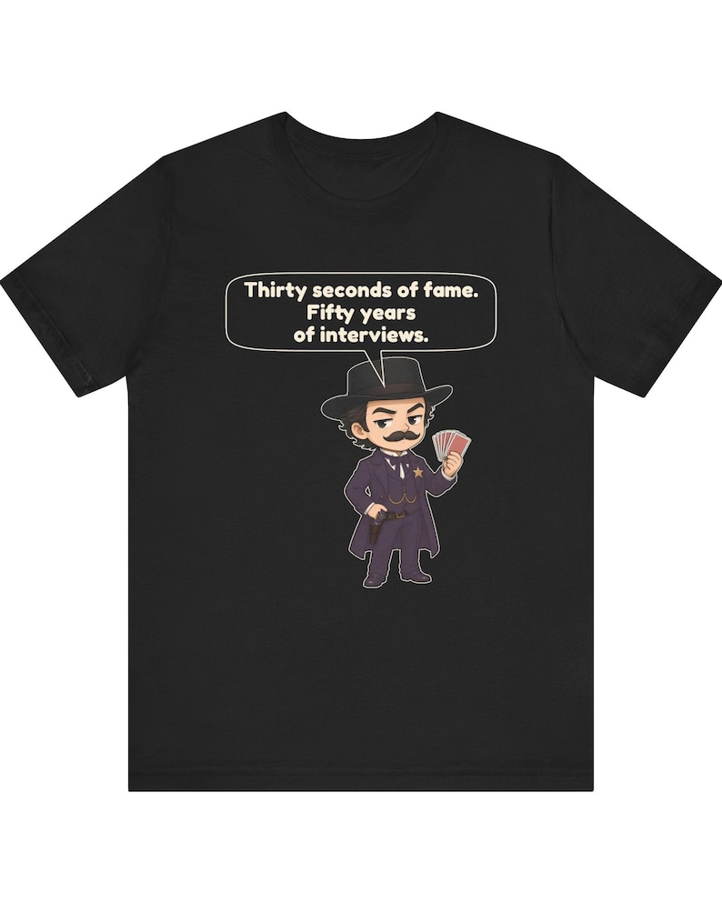 Pu&ograve; includere: Una t-shirt nera con un'illustrazione in stile cartoon di un personaggio con i baffi, cappotto scuro e cappello che tiene in mano delle carte da gioco. Un fumetto sopra il personaggio contiene il testo: Thirty seconds of fame. Fifty years of interviews.