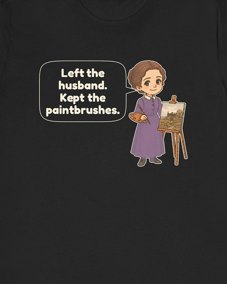 May include: A black t-shirt featuring a cartoon illustration of a person in a purple dress holding a paintbrush and palette next to an easel. A speech bubble contains the text: Left the husband. Kept the paintbrushes.