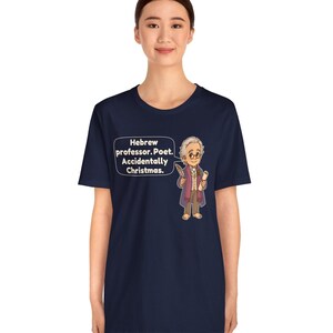 Pu&ograve; includere: Una t-shirt blu navy a maniche corte con un'illustrazione a cartone animato di un uomo anziano con gli occhiali che tiene in mano una penna e una pergamena. Un fumetto accanto al personaggio contiene il testo: Hebrew professor. Poet. Accidentally Christmas.