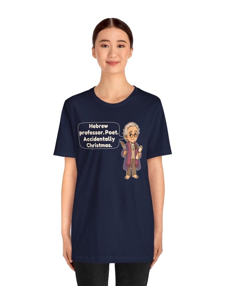 Pu&ograve; includere: Una t-shirt blu navy a maniche corte con un'illustrazione a cartone animato di un uomo anziano con gli occhiali che tiene in mano una penna e una pergamena. Un fumetto accanto al personaggio contiene il testo: Hebrew professor. Poet. Accidentally Christmas.