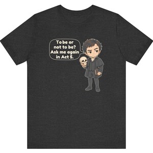 以下が含まれることがあります： ダークヘザーグレーのTシャツ。歴史的な衣装を着た人物が頭蓋骨を持っている漫画風のイラストが描かれています。吹き出しには「To be or not to be? Ask me again in Act 5.」というテキストが含まれています。デザインは胸の中央に配置されています。