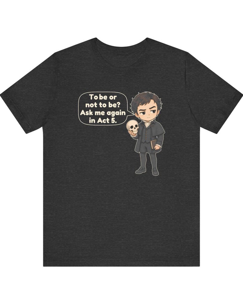 以下が含まれることがあります： ダークヘザーグレーのTシャツ。歴史的な衣装を着た人物が頭蓋骨を持っている漫画風のイラストが描かれています。吹き出しには「To be or not to be? Ask me again in Act 5.」というテキストが含まれています。デザインは胸の中央に配置されています。