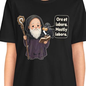 May include: A black t-shirt featuring a cartoon monk with a white beard, holding a wooden staff and an open book. A black crow sits on the book holding a piece of bread. A speech bubble contains the text: Ora et labora. Mostly labora.