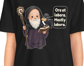 May include: A black t-shirt featuring a cartoon monk with a white beard, holding a wooden staff and an open book. A black crow sits on the book holding a piece of bread. A speech bubble contains the text: Ora et labora. Mostly labora.