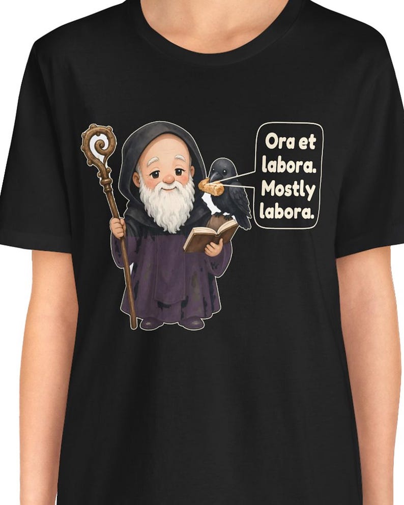 May include: A black t-shirt featuring a cartoon monk with a white beard, holding a wooden staff and an open book. A black crow sits on the book holding a piece of bread. A speech bubble contains the text: Ora et labora. Mostly labora.