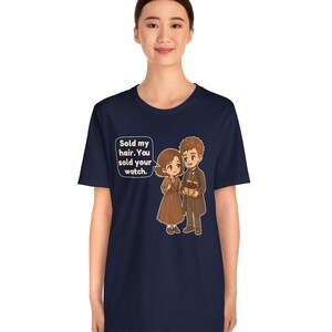 May include: A navy blue short-sleeve t-shirt featuring a cartoon illustration of two figures in brown clothing. A speech bubble contains the text: Sold my hair. You sold your watch. The design is centered on the front of the garment.