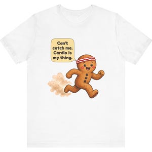 May include: A white t-shirt featuring a cartoon gingerbread person wearing a red and white striped headband, running and leaving a dust cloud behind. A speech bubble above contains the text: Can't catch me. Cardio is my thing.