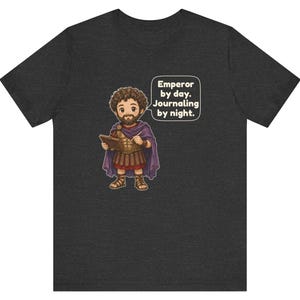 K&ouml;nnte beinhalten: Ein dunkelgrau meliertes T-Shirt mit einer Cartoon-Illustration eines b&auml;rtigen Kaisers in r&ouml;mischer R&uuml;stung und violettem Umhang, der ein Tagebuch und einen Stift h&auml;lt. Eine Sprechblase enth&auml;lt den Text: Emperor by day. Journaling by night.