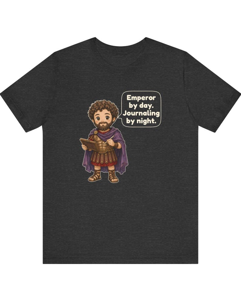 K&ouml;nnte beinhalten: Ein dunkelgrau meliertes T-Shirt mit einer Cartoon-Illustration eines b&auml;rtigen Kaisers in r&ouml;mischer R&uuml;stung und violettem Umhang, der ein Tagebuch und einen Stift h&auml;lt. Eine Sprechblase enth&auml;lt den Text: Emperor by day. Journaling by night.