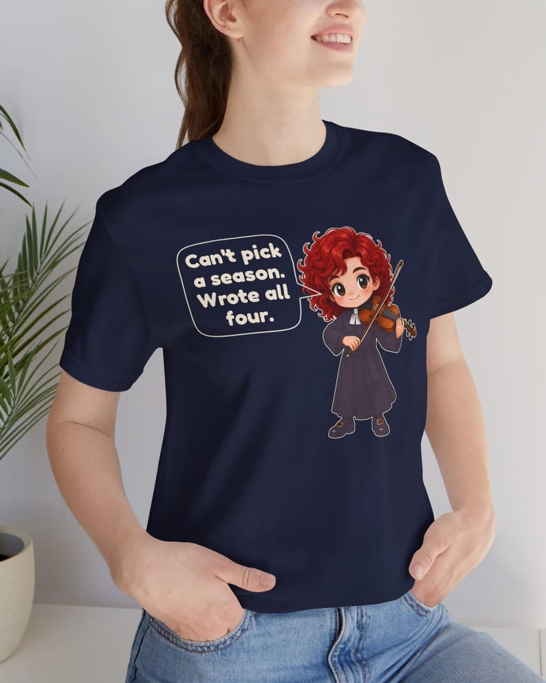 May include: A navy blue t-shirt featuring a cartoon illustration of a red-haired character playing a violin. A speech bubble contains the text: Can't pick a season. Wrote all four. The character wears a dark robe with a white collar.