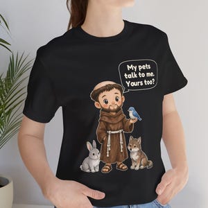 May include: A black t-shirt featuring a cartoon illustration of a monk in a brown robe standing with a grey rabbit, a small brown dog, and a blue bird. A speech bubble above the monk contains the text: My pets talk to me. Yours too?