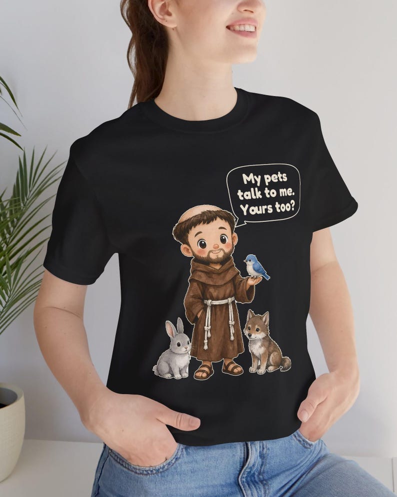 May include: A black t-shirt featuring a cartoon illustration of a monk in a brown robe standing with a grey rabbit, a small brown dog, and a blue bird. A speech bubble above the monk contains the text: My pets talk to me. Yours too?