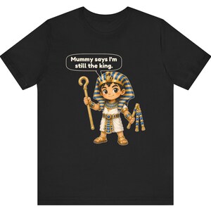 Może przedstawiać: Czarny t-shirt z rysunkową ilustracją młodego faraona w niebiesko-złotym nakryciu głowy, białej tunice i sandałach. Postać trzyma złote berło i bicz, a nad nią znajduje się dymek z napisem Mummy says I'm still the king.