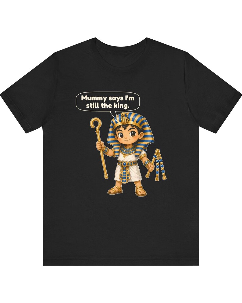 Może przedstawiać: Czarny t-shirt z rysunkową ilustracją młodego faraona w niebiesko-złotym nakryciu głowy, białej tunice i sandałach. Postać trzyma złote berło i bicz, a nad nią znajduje się dymek z napisem Mummy says I'm still the king.