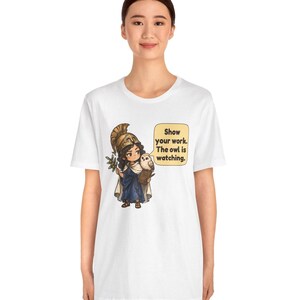 Puede incluir: Una camiseta blanca con un gr&aacute;fico de un personaje de dibujos animados con casco dorado y t&uacute;nica azul, sosteniendo una rama de olivo y un peque&ntilde;o b&uacute;ho. Un bocadillo contiene el texto: Show your work. The owl is watching.