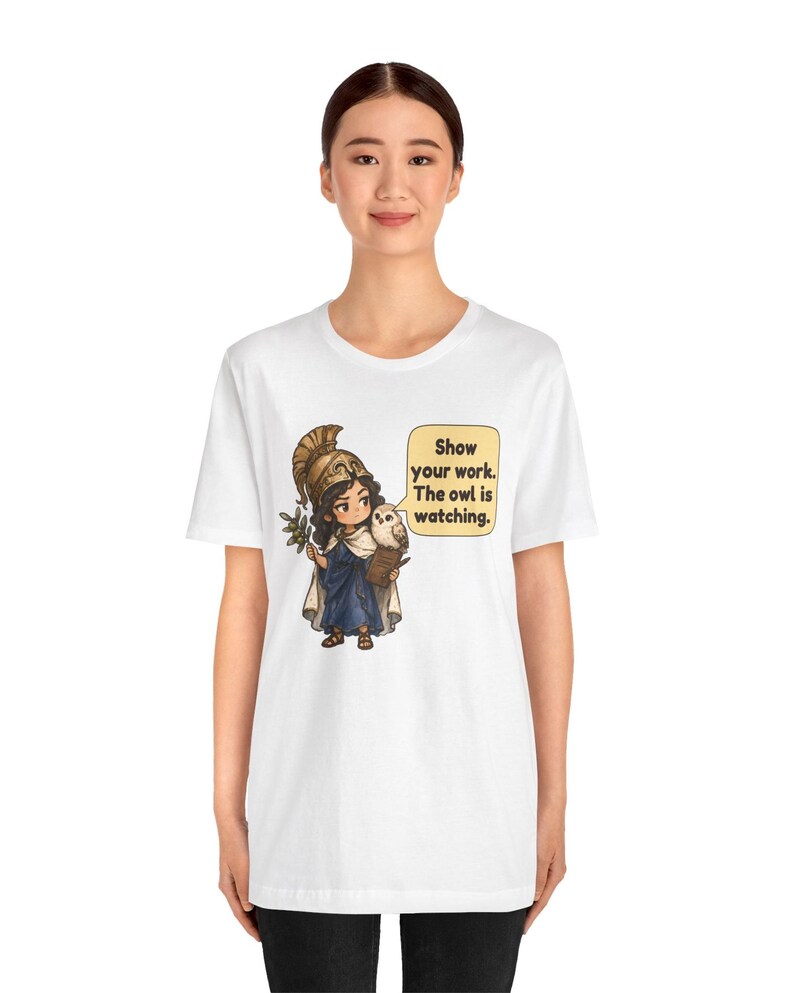 Puede incluir: Una camiseta blanca con un gr&aacute;fico de un personaje de dibujos animados con casco dorado y t&uacute;nica azul, sosteniendo una rama de olivo y un peque&ntilde;o b&uacute;ho. Un bocadillo contiene el texto: Show your work. The owl is watching.