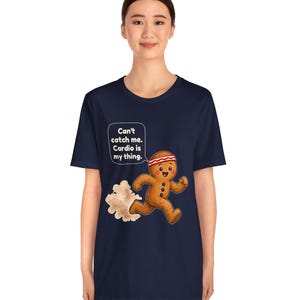 May include: A navy blue t-shirt featuring a graphic of a running gingerbread person wearing a red and white headband. A speech bubble above the character contains the text: Can't catch me. Cardio is my thing. A cloud of dust trails behind the figure.