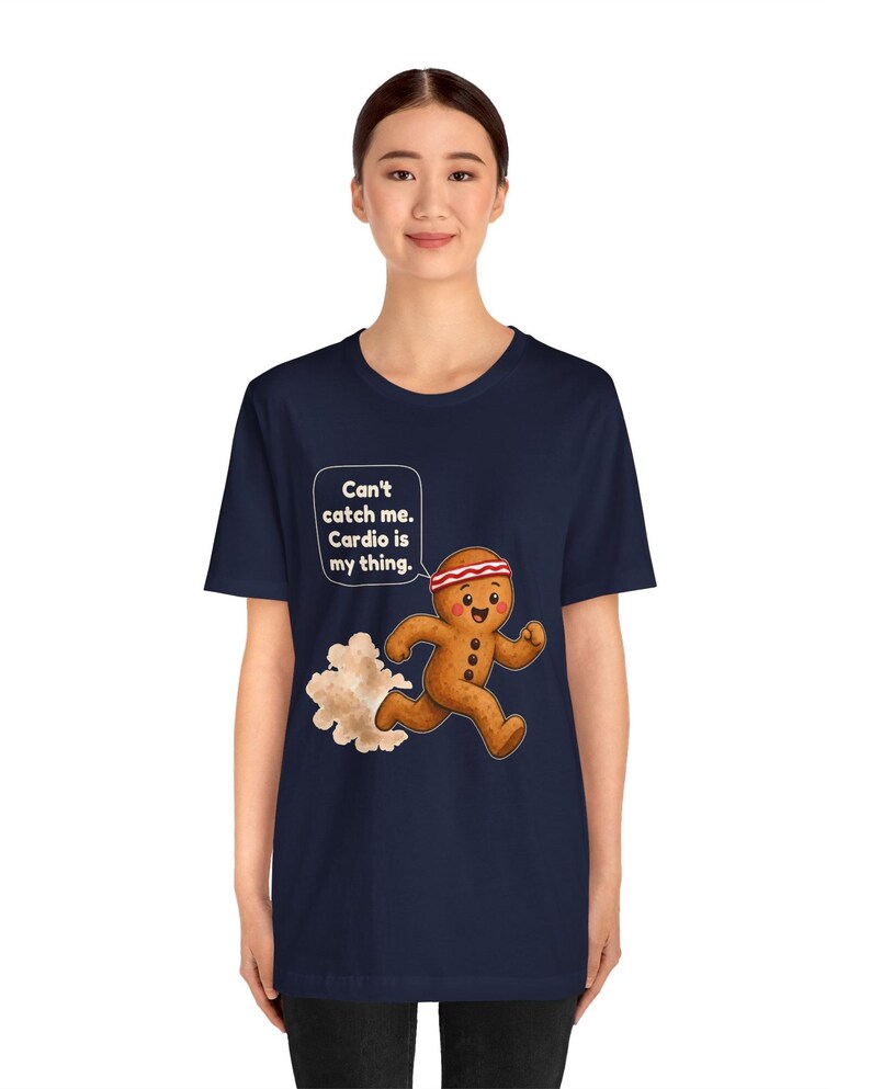 May include: A navy blue t-shirt featuring a graphic of a running gingerbread person wearing a red and white headband. A speech bubble above the character contains the text: Can't catch me. Cardio is my thing. A cloud of dust trails behind the figure.