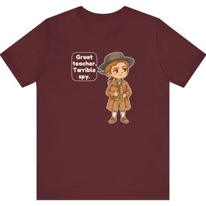 May include: A maroon short-sleeved t-shirt featuring a cartoon illustration of a person in 18th-century attire, including a hat and coat, holding a book. A speech bubble next to the character contains the text: Great teacher. Terrible spy.
