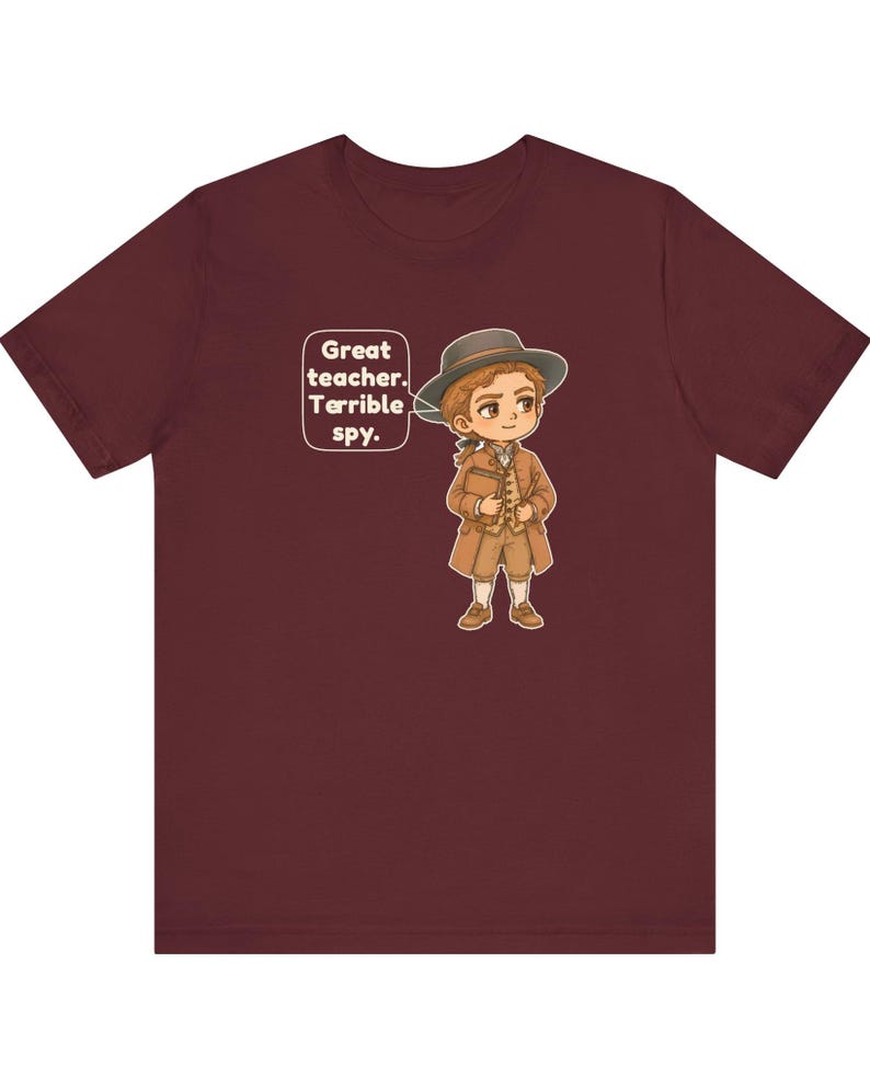 May include: A maroon short-sleeved t-shirt featuring a cartoon illustration of a person in 18th-century attire, including a hat and coat, holding a book. A speech bubble next to the character contains the text: Great teacher. Terrible spy.