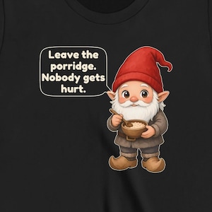 May include: A black t-shirt featuring a graphic of a gnome wearing a red hat and brown clothing, holding a wooden bowl and spoon. A speech bubble next to the gnome contains the text: Leave the porridge. Nobody gets hurt.