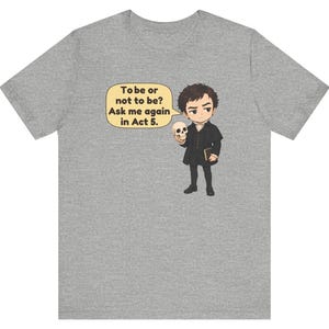 以下が含まれることがあります： ヘザーグレーのTシャツ。暗い色の服を着た人物が頭蓋骨と本を持っている漫画風のイラストが描かれています。吹き出しには「To be or not to be? Ask me again in Act 5.」というテキストが含まれています。デザインは中央に配置されています。