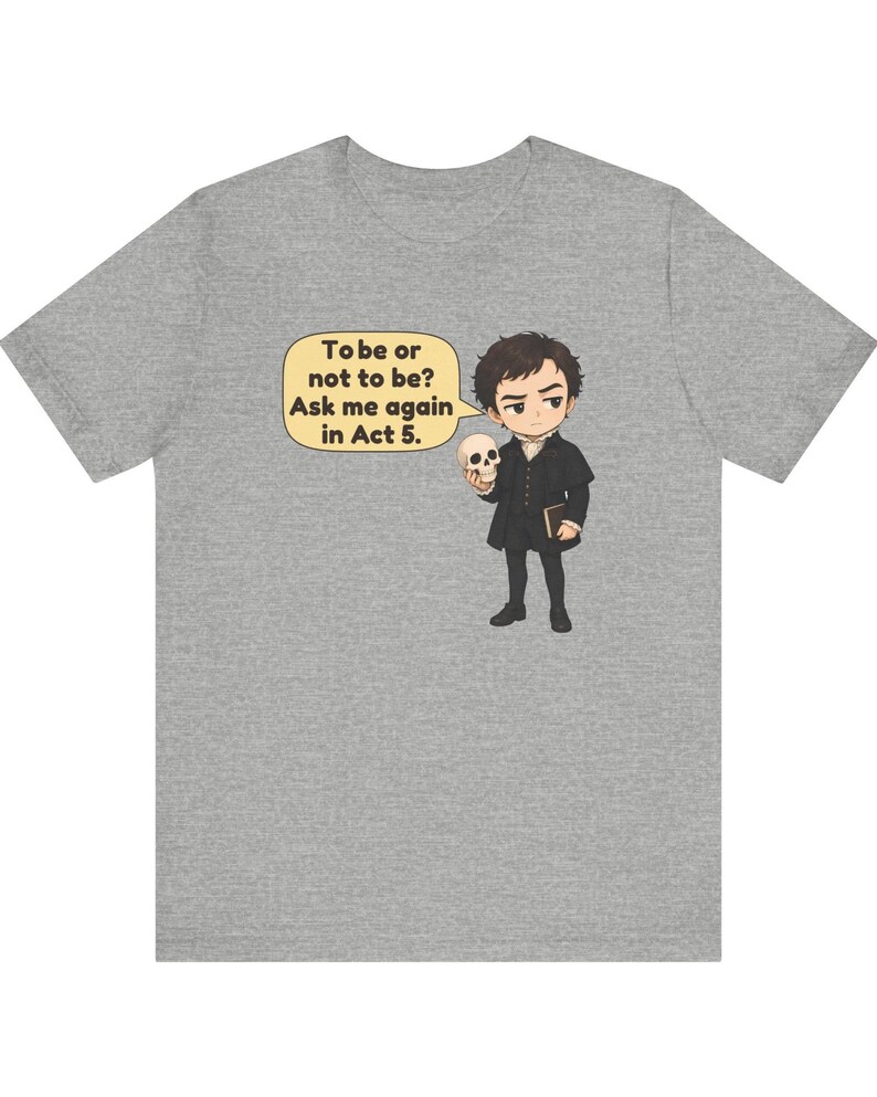 以下が含まれることがあります： ヘザーグレーのTシャツ。暗い色の服を着た人物が頭蓋骨と本を持っている漫画風のイラストが描かれています。吹き出しには「To be or not to be? Ask me again in Act 5.」というテキストが含まれています。デザインは中央に配置されています。