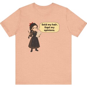May include: A peach-colored t-shirt featuring a cartoon illustration of a person with dark braided hair and a red bow, wearing a brown dress and apron. The character holds a quill and paper, with a speech bubble stating: Sold my hair. Kept my opinions.