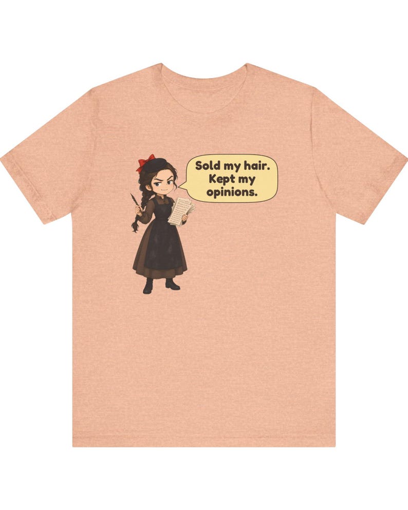 May include: A peach-colored t-shirt featuring a cartoon illustration of a person with dark braided hair and a red bow, wearing a brown dress and apron. The character holds a quill and paper, with a speech bubble stating: Sold my hair. Kept my opinions.
