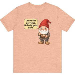 May include: A peach-coloured t-shirt featuring a graphic of a gnome wearing a red hat and brown clothing, holding a bowl of porridge. A speech bubble above the gnome contains the text: Leave the porridge. Nobody gets hurt.