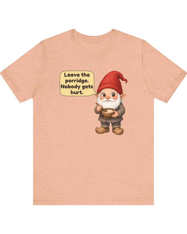 May include: A peach-coloured t-shirt featuring a graphic of a gnome wearing a red hat and brown clothing, holding a bowl of porridge. A speech bubble above the gnome contains the text: Leave the porridge. Nobody gets hurt.