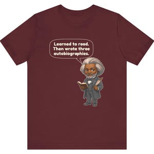 Puede incluir: Una camiseta de manga corta color granate con una ilustraci&oacute;n de dibujos animados de un hombre con pelo gris y barba, vestido con traje, sosteniendo un libro y una pluma. Un bocadillo sobre la figura contiene el texto: Learned to read. Then wrote three autobiographies.