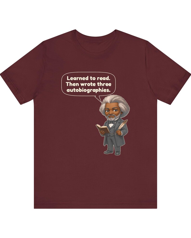 Puede incluir: Una camiseta de manga corta color granate con una ilustraci&oacute;n de dibujos animados de un hombre con pelo gris y barba, vestido con traje, sosteniendo un libro y una pluma. Un bocadillo sobre la figura contiene el texto: Learned to read. Then wrote three autobiographies.
