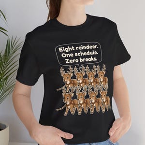 K&ouml;nnte beinhalten: Ein schwarzes T-Shirt mit einer Grafik von acht braunen Rentieren, die in zwei Reihen angeordnet sind. &Uuml;ber den Rentieren befindet sich eine wei&szlig;e Sprechblase mit dem Text: Eight reindeer. One schedule. Zero breaks. Das Motiv ist mittig.
