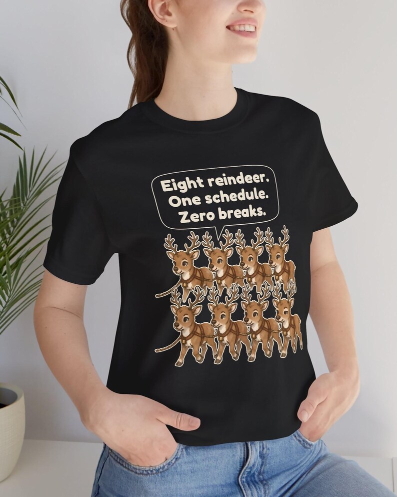 K&ouml;nnte beinhalten: Ein schwarzes T-Shirt mit einer Grafik von acht braunen Rentieren, die in zwei Reihen angeordnet sind. &Uuml;ber den Rentieren befindet sich eine wei&szlig;e Sprechblase mit dem Text: Eight reindeer. One schedule. Zero breaks. Das Motiv ist mittig.