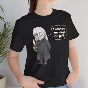 May include: A black t-shirt featuring a graphic illustration of a pale, long-haired vampire in a dark coat holding a lit candle. A speech bubble next to the character contains the text: I don't do mornings. Or garlic.