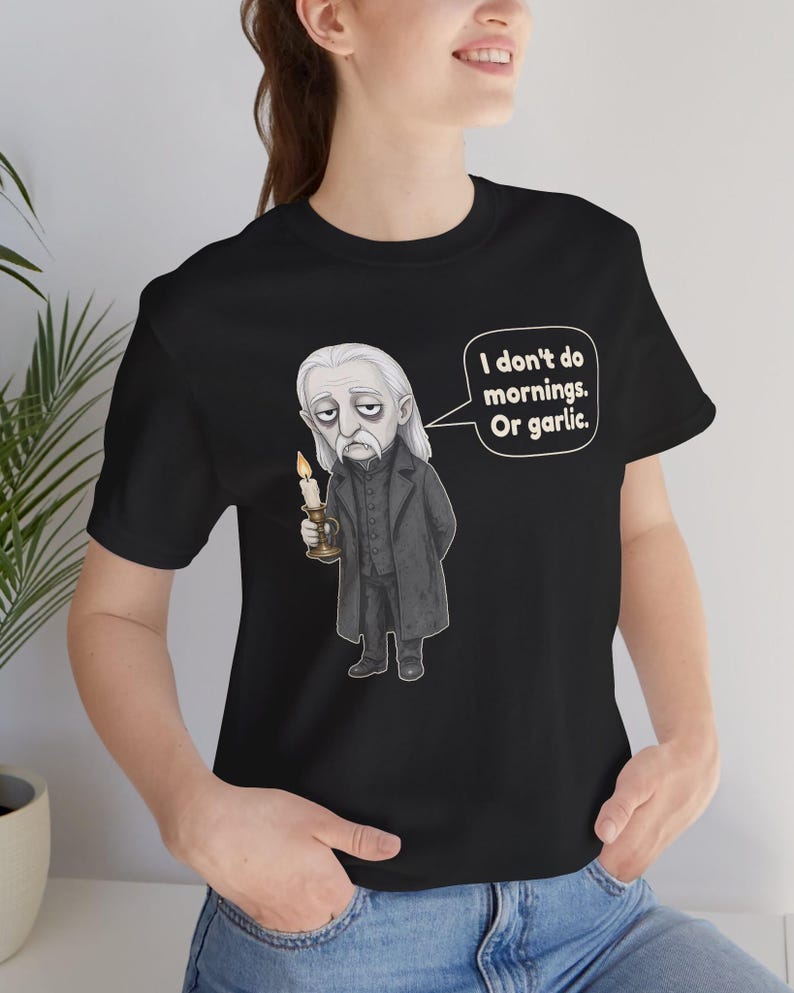 May include: A black t-shirt featuring a graphic illustration of a pale, long-haired vampire in a dark coat holding a lit candle. A speech bubble next to the character contains the text: I don't do mornings. Or garlic.