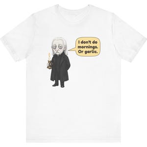 May include: A white short-sleeved t-shirt featuring a graphic of a pale character in a black coat holding a lit candle. A speech bubble next to the character contains the text: I don't do mornings. Or garlic. The design is centered on the chest.