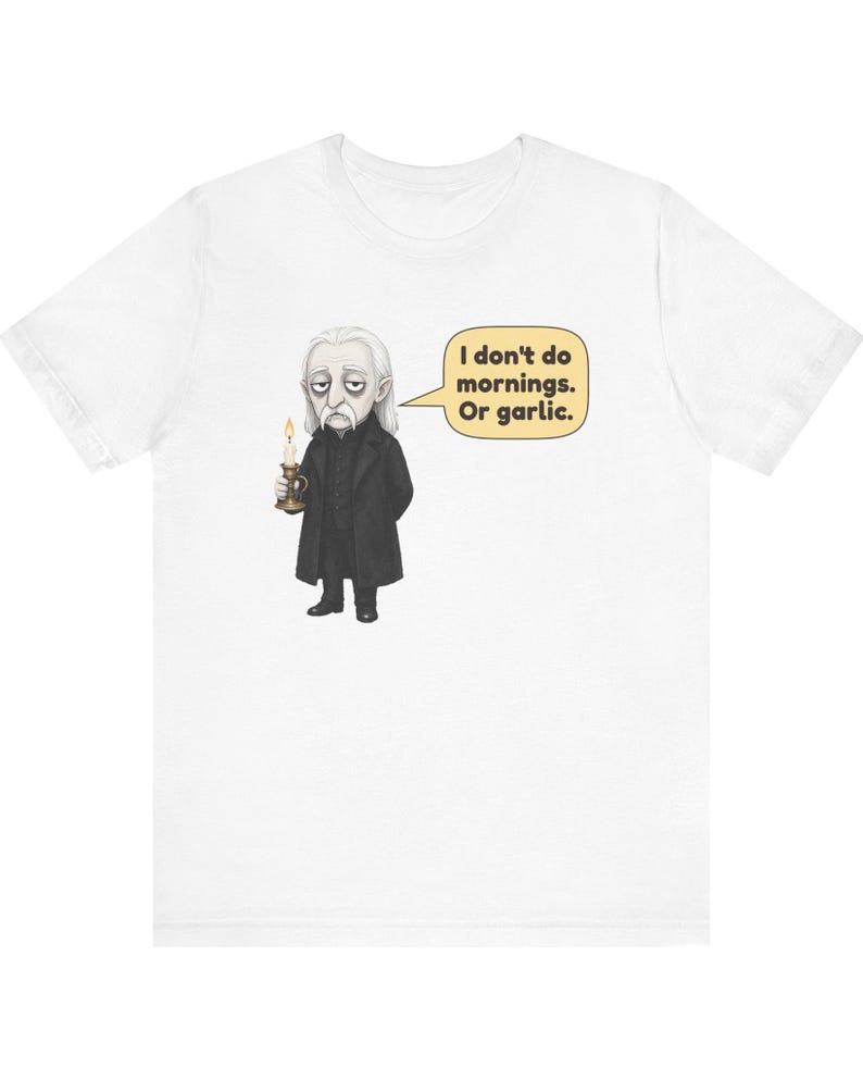 May include: A white short-sleeved t-shirt featuring a graphic of a pale character in a black coat holding a lit candle. A speech bubble next to the character contains the text: I don't do mornings. Or garlic. The design is centered on the chest.