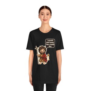 May include: A black short-sleeve t-shirt featuring a cartoon illustration of a bearded figure holding a wooden staff and a honey dipper. A speech bubble above the figure contains the text: Locusts and honey. Very trendy diet.