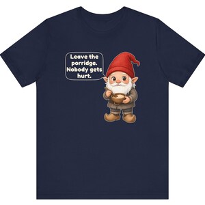 May include: A navy blue short-sleeve t-shirt featuring a graphic of a gnome wearing a red hat and brown clothing, holding a bowl of porridge with a spoon. A speech bubble next to the gnome contains the text: Leave the porridge. Nobody gets hurt.