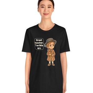 May include: A black t-shirt featuring a cartoon illustration of a person wearing a brown coat, vest, and hat. A speech bubble next to the character contains the text: Great teacher. Terrible spy. The design is centered on the chest area.