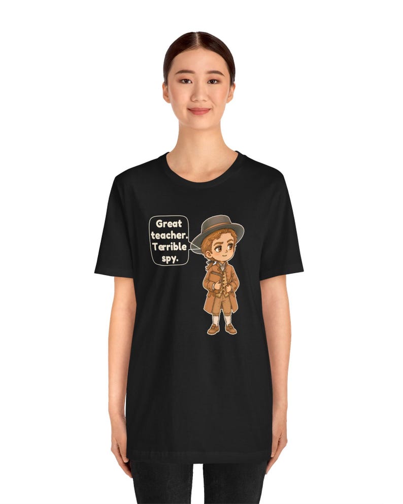 May include: A black t-shirt featuring a cartoon illustration of a person wearing a brown coat, vest, and hat. A speech bubble next to the character contains the text: Great teacher. Terrible spy. The design is centered on the chest area.