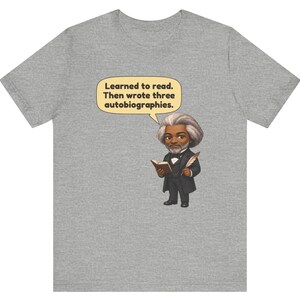 Puede incluir: Una camiseta gris jaspeado con una ilustraci&oacute;n de dibujos animados de Frederick Douglass sosteniendo un libro y una pluma. Un globo de texto amarillo sobre la figura contiene el texto: Learned to read. Then wrote three autobiographies.