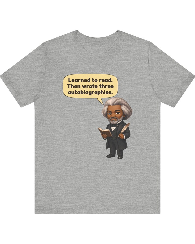 Puede incluir: Una camiseta gris jaspeado con una ilustraci&oacute;n de dibujos animados de Frederick Douglass sosteniendo un libro y una pluma. Un globo de texto amarillo sobre la figura contiene el texto: Learned to read. Then wrote three autobiographies.